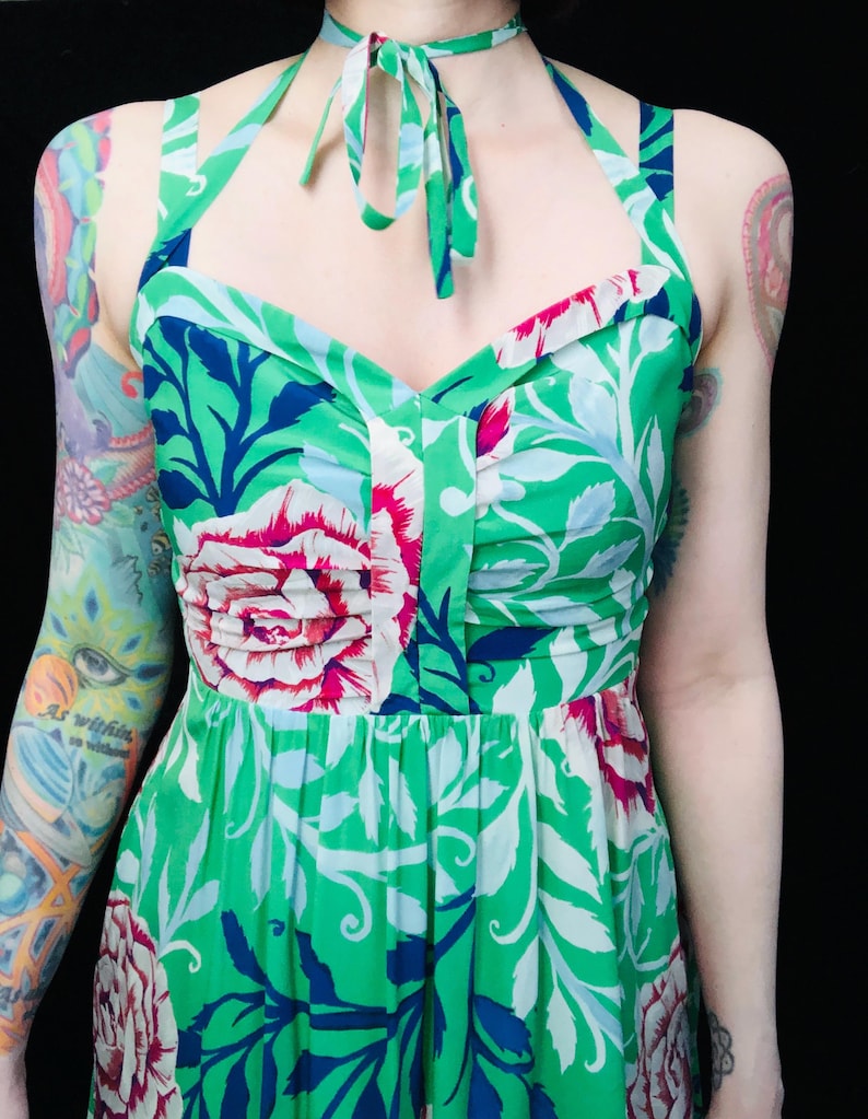 May include: A green halter dress with a floral pattern featuring large pink and white flowers, blue accents, and white leaves. The dress has a tie at the neck and a fitted bodice. The fabric is a vibrant green.