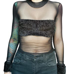 May include: Black mesh top with long sleeves, a mock neck, and a fuzzy chest band. The cuffs also feature fuzzy trim. The top is paired with dark wash jeans.