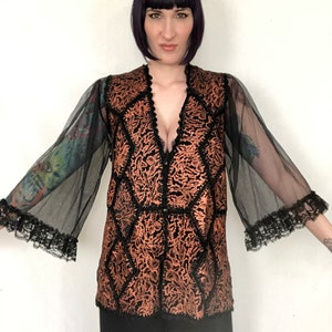 Puede incluir: Una blusa negra transparente con un diseño de brocado de color cobre y ribetes de encaje en las mangas. La blusa tiene un escote en V profundo y un corte holgado.