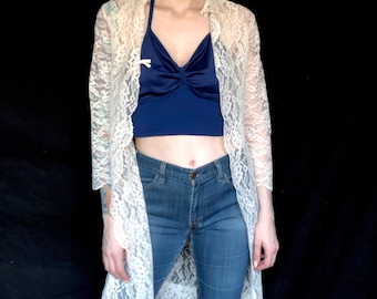 May include: A white lace long-sleeved cardigan with a blue satin halter top underneath. The person is wearing blue jeans.