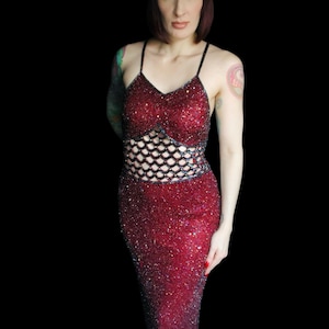 May include: A sparkling, red sequined gown with a fitted bodice and a fish-tail skirt. The dress features a black net-like design at the waist and thin spaghetti straps. The dress has a high slit in the front.