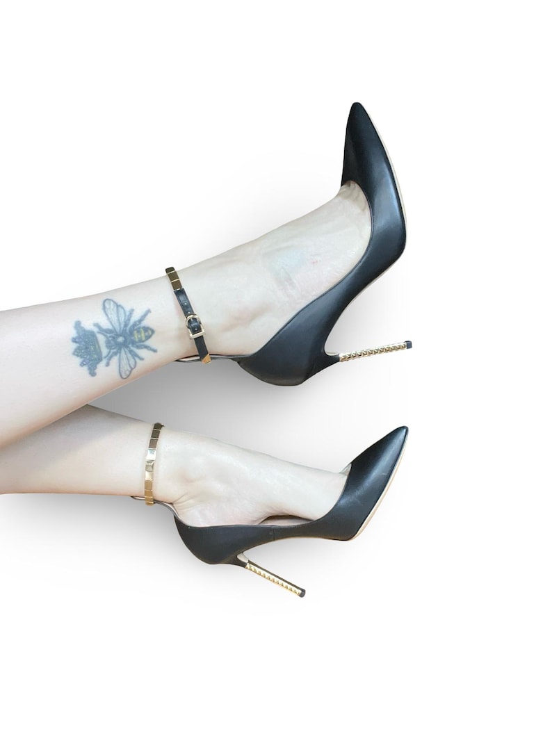 May include: Black leather high heels with pointed toes and gold-colored stiletto heels. The shoes have ankle straps with gold-colored buckles. A tattoo of a bee is visible on the ankle.