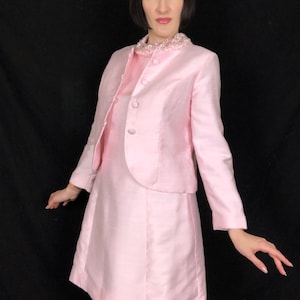 May include: A pink dress suit with a beaded collar and a fitted jacket. The dress has a simple A-line silhouette and the jacket has a classic collar and button closure.