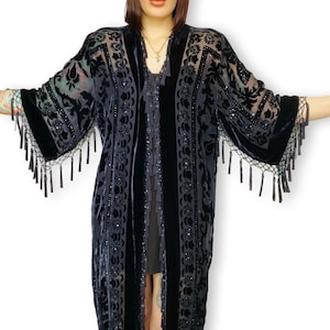 May include: A long, dark blue velvet kimono-style jacket with a floral pattern and fringe trim. The jacket has wide sleeves and is embellished with sequins. The garment is open in the front and drapes to the ankles.
