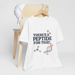 May include: White t-shirt with the text "THERE'S A PEPTIDE FOR THAT" in dark blue and brown. The shirt features a molecular structure and a DNA strand graphic. A white baseball cap rests on a wooden chair.