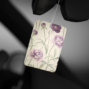 Op de afbeelding: Een witte autoluchtverfrisser met een bloemenpatroon. Het patroon bevat paarse en roze bloemen met groene stelen en bladeren.