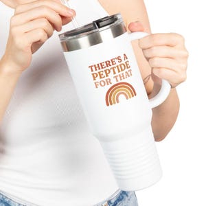 Puede incluir: Taza de viaje blanca con asa y tapa negra. La taza presenta el texto "THERE'S A PEPTIDE FOR THAT" sobre un gráfico de arcoíris marrón y naranja. La taza está diseñada para contener bebidas.