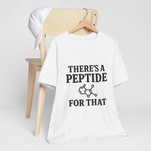 May include: White t-shirt with the text "THERE'S A PEPTIDE FOR THAT" in black, along with a molecular structure graphic. The shirt is hanging on a wooden hanger, with a white cap resting on a wooden chair.