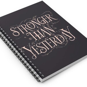 Puede incluir: Una libreta espiral negra con el texto "Stronger Than Yesterday" en blanco, en una fuente decorativa. El texto está rodeado por un diseño floral de color rosa claro.