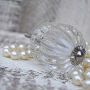 May include: A clear glass cabinet knob with a ribbed design and a silver-colored base. The knob is lying on a white surface next to a strand of cream-colored pearls. The overall theme is vintage decor.