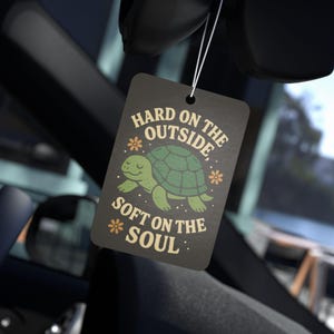 Puede incluir: Ambientador de coche negro con una ilustración de tortuga verde y el texto "Hard on the outside, soft on the soul."