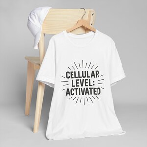Puede incluir: Camiseta blanca con el texto negro "CELLULAR LEVEL: ACTIVATED", colgada de una percha de madera. Una gorra de béisbol blanca descansa sobre el respaldo de una silla de madera clara. La camiseta es el punto focal.