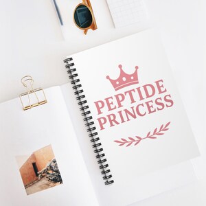 Może przedstawiać: Biały notatnik w spiralę z różowym napisem "PEPTIDE PRINCESS", zwieńczony koroną i gałązką liści. Widoczny jest również złoty spinacz i okulary przeciwsłoneczne.