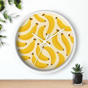 May include: White-framed wall clock with a yellow banana pattern on a cream background. The clock hands are white. The clock is on a wooden surface with a small potted plant to the right and green leaves to the left.