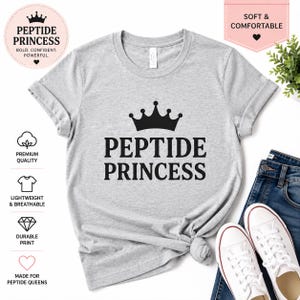 May include: A light gray t-shirt with the words "PEPTIDE PRINCESS" in bold black letters, topped with a black crown graphic. The shirt is paired with blue jeans and white sneakers. The shirt also has the text "SOFT & COMFORTABLE".