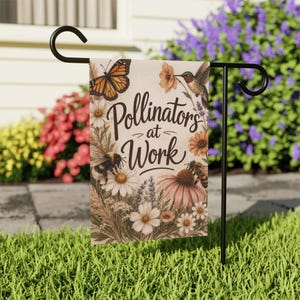 Puede incluir: Una bandera de jardín con una mariposa, un colibrí y abejas entre flores. La bandera dice "Pollinators at Work". El diseño incluye margaritas, equináceas y otras flores silvestres, sobre un fondo neutro. La bandera está en un soporte de metal negro.