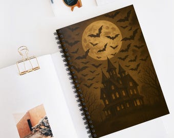 May include: A spiral-bound notebook featuring a spooky Halloween design. The cover depicts a haunted house with a full moon, bats, and bare trees in shades of brown and black. The notebook is open on a white surface.