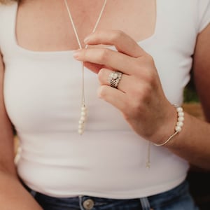 May include: A woman wearing a white tank top and blue jeans is holding a silver chain necklace with a pearl drop. She is also wearing a silver ring with a large stone on her ring finger and a pearl bracelet on her wrist.