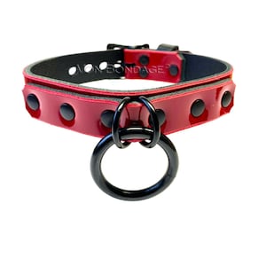 May include: A red patent leather collar with black hardware. The collar features a large black metal ring and black studs. The text "VON BONDAGE" is visible on the collar.