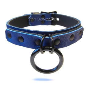 May include: A blue leather collar with black hardware, including a large ring and buckle. The collar is riveted and features the text "Von Bondage".