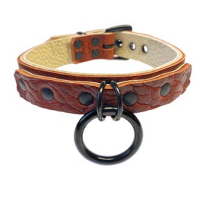 May include: A brown leather collar with a textured pattern and black hardware. The collar features a large black metal ring and black rivets. The interior of the collar is a light beige color. The collar is designed for pets.