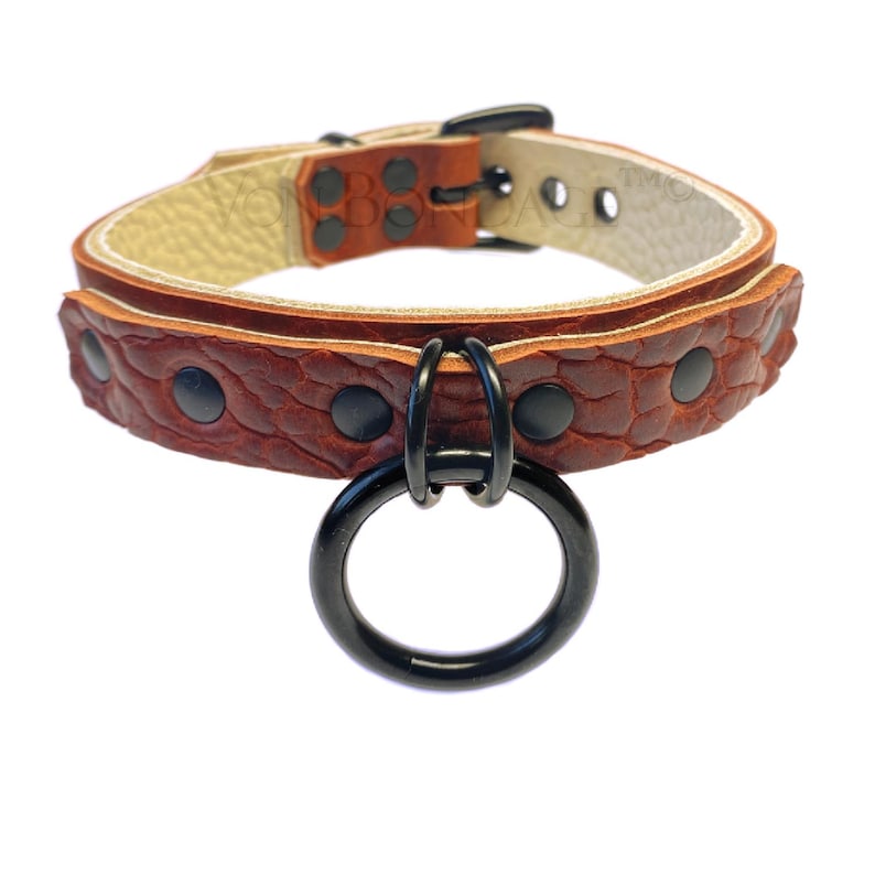 May include: A brown leather collar with a textured pattern and black hardware. The collar features a large black metal ring and black rivets. The interior of the collar is a light beige color. The collar is designed for pets.