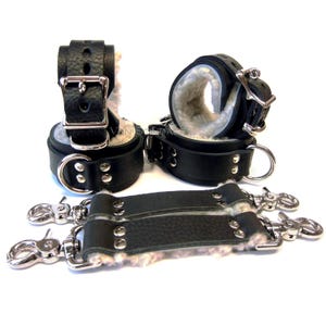 May include: Black leather wrist cuffs with silver hardware and a soft white fleece lining. The cuffs are connected by two black leather straps with silver clasps.