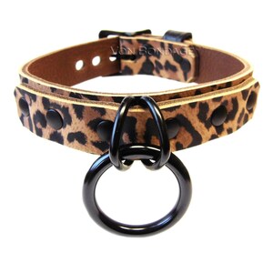 May include: A leopard print collar with a black metal ring and hardware. The collar is made of brown leather and features black rivets. The text "VON BONDAGE" is visible on the collar.