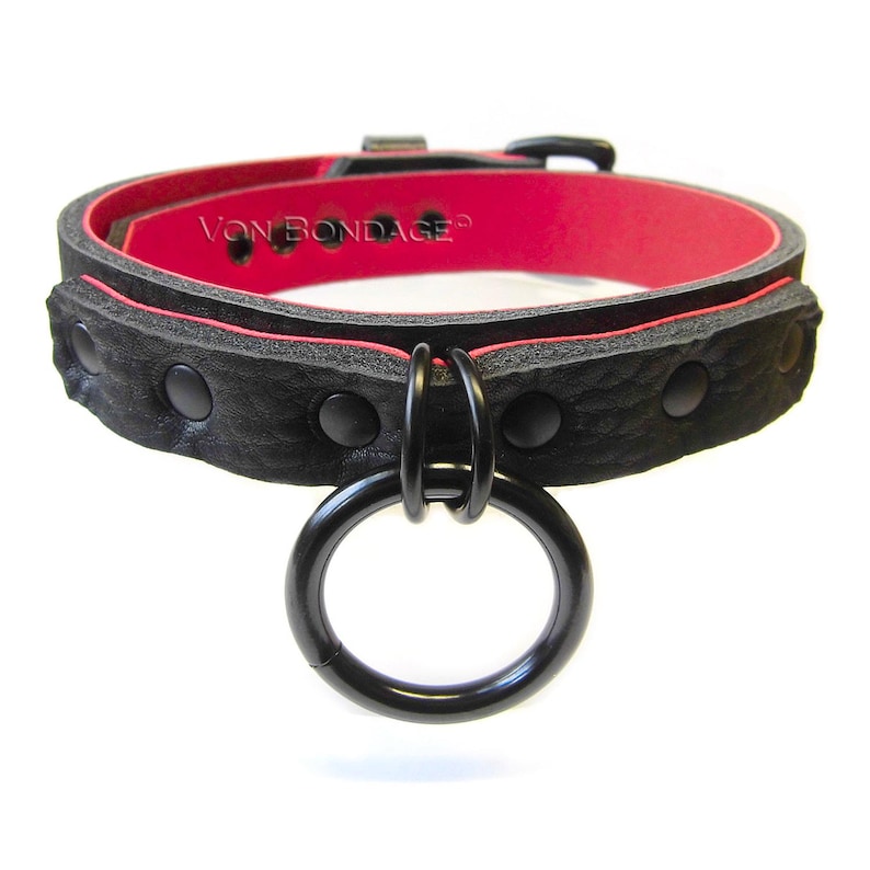 May include: Black leather collar with a red interior and a large black metal ring. The collar has black rivets and the text "Von Bondage".
