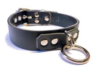 May include: A dark gray leather choker with silver-toned hardware and a large, round, silver-toned ring attached. The choker has multiple silver-toned studs.