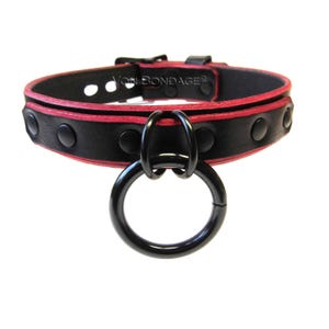 May include: Black leather collar with red trim and a large black metal ring. The collar is secured with a buckle and features black studs. The text "Von Bondage" is visible on the collar.