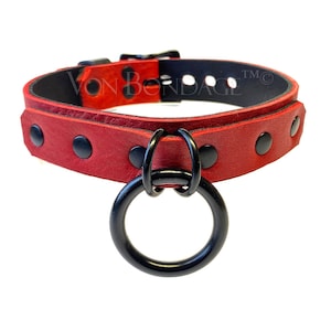 May include: Red leather collar with black hardware, including a large O-ring and rivets. The collar has a buckle closure and is designed to be worn around the neck. The brand name "VON BONDAGE" is visible on the collar.