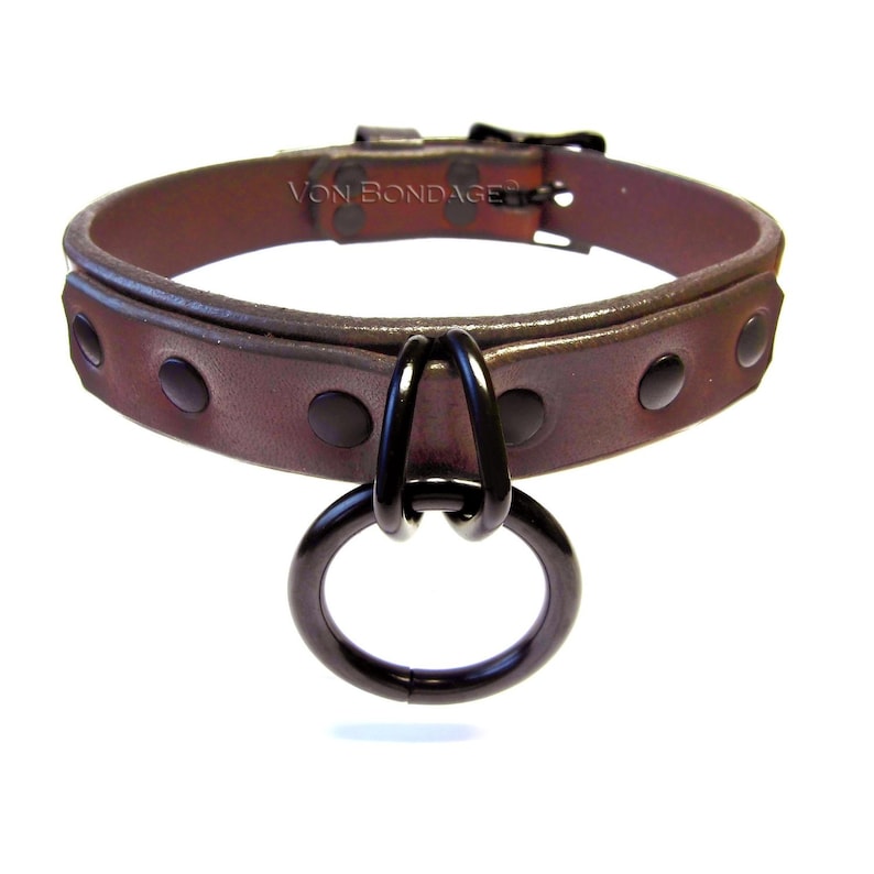 May include: Brown leather collar with black metal hardware, including a large O-ring and rivets. The collar has the text "Von BONDAGE" on the top. The collar is designed to be worn around the neck.