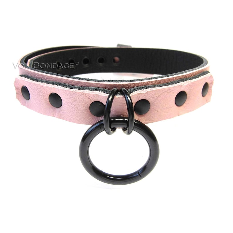 May include: A pink leather collar with black rivets and a large black metal ring. The collar has a black interior and is secured with a buckle. The text "Von Bondage" is visible on the left side of the collar.