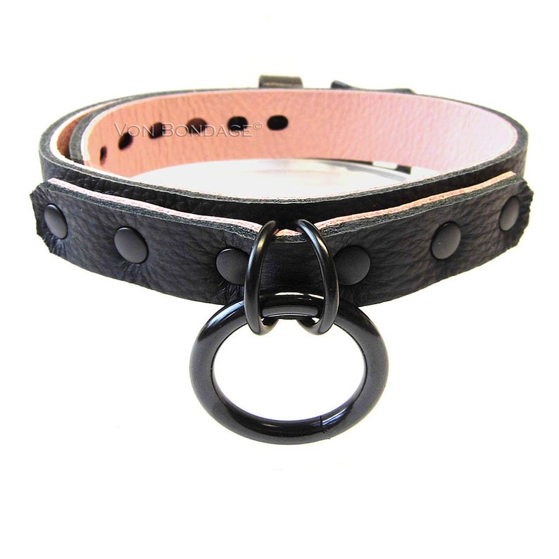 May include: Black leather collar with a pink interior and a large black metal ring. The collar is secured with black rivets and features the text "Von Bondage".