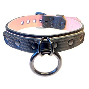 May include: Black leather collar with a pink interior and a large metal ring. The collar has black metal studs and a buckle closure. The text "IN BONDAGE" is visible on the interior of the collar.