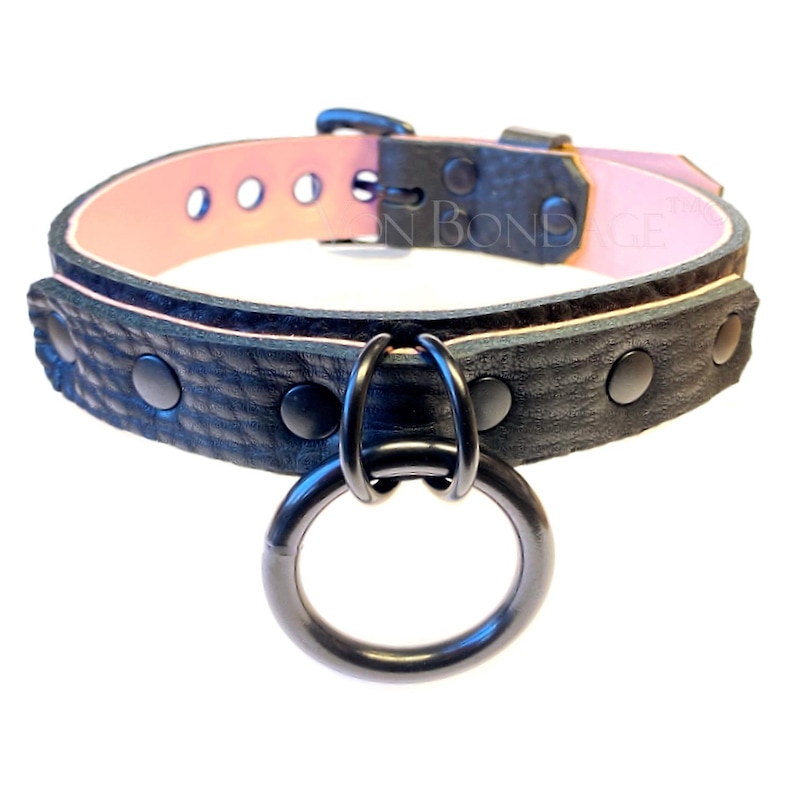 May include: Black leather collar with a pink interior and a large metal ring. The collar has black metal studs and a buckle closure. The text "IN BONDAGE" is visible on the interior of the collar.