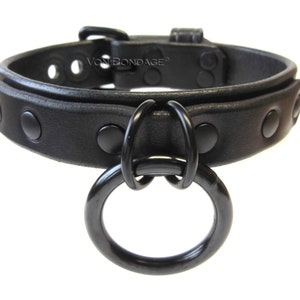 May include: A black leather collar with a large, round, black metal ring attached. The collar has a buckle closure and is adorned with evenly spaced, black rivets. The text "Von Bondage" is visible on the collar.