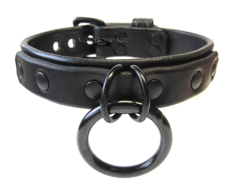 May include: A black leather collar with a large, round, black metal ring attached. The collar has a buckle closure and is adorned with evenly spaced, black rivets. The text "Von Bondage" is visible on the collar.
