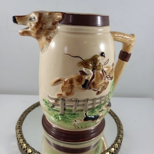 Puede incluir: Una jarra de cerámica con un caño en forma de cabeza de zorro, que representa a un cazador a caballo con perros que persiguen a un zorro. La jarra tiene un asa marrón y una base marrón y crema.