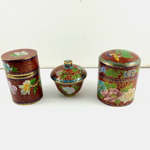 Puede incluir: Tres pequeñas cajas redondas de cloisonné rojo, dorado y azul con diseños florales. Las cajas son de metal y tienen un acabado brillante.