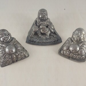May include: Three silver-toned metal figurines of a smiling Buddha. The figurines are detailed with intricate carvings and are in various poses.