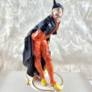 May include: A porcelain figurine of a man in period costume. The figure is dressed in a black coat, orange breeches, and yellow shoes. The figurine has a black hat and a beard. The base is white with gold detailing.
