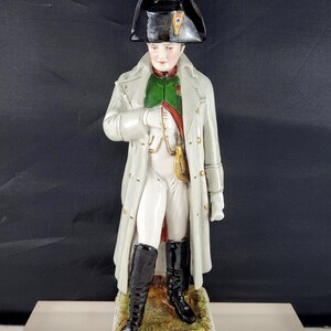 Puede incluir: Figura de porcelana de Napoleón Bonaparte, vestido con un abrigo blanco, botas negras y un sombrero bicornio. La figura se alza sobre una base rectangular con ribetes dorados y la inscripción "Napoleón".