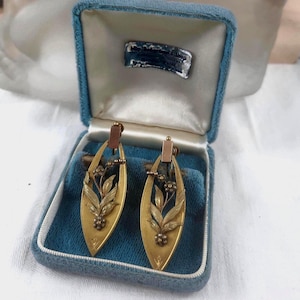 May include: A pair of vintage gold-toned earrings with a floral design, presented in a blue velvet box. The earrings have a long, oval shape with detailed leaf and flower accents. The box has a white satin lining and a small label.
