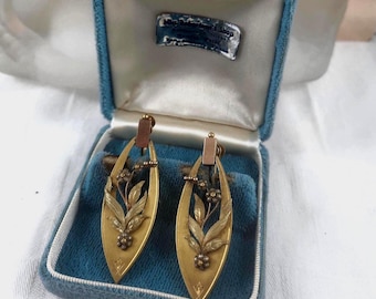May include: A pair of vintage gold-toned earrings with a floral design, presented in a blue velvet box. The earrings have a long, oval shape with detailed leaf and flower accents. The box has a white satin lining and a small label.