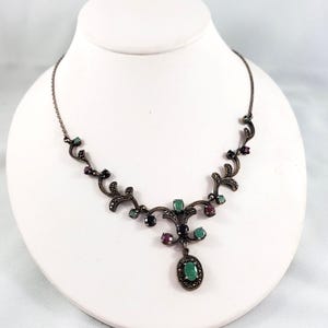 Op de afbeelding: Een ketting van donker metaal met een reeks versierde, rankachtige schakels bezet met kleine, ronde, donkergrijze stenen. Het ontwerp is geaccentueerd met ovaal geslepen groene en dieprode edelstenen en een centrale ovale groene hanger.
