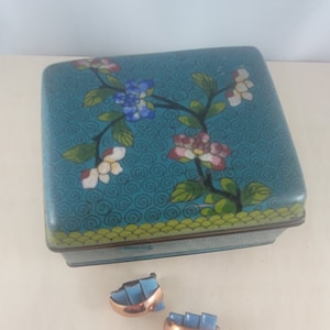 May include: A teal blue square box with a floral design and a pair of blue and copper earrings.