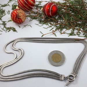 May include: A silver chain necklace with a toggle clasp. The chain is made of three strands of silver, each with a herringbone pattern. The necklace is lying on a white surface.