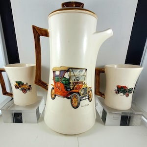 May include: A vintage cream-colored coffee pot and two matching mugs. The coffee pot and mugs feature a brown handle and a colorful illustration of a Ford T 1908 car. The coffee pot has a brown lid and spout.